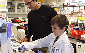 Photo of a man wearing a black sweater and a boy wearing a white lab coat; they are conducting a lab experiment by putting algae from a pipette onto a slide.