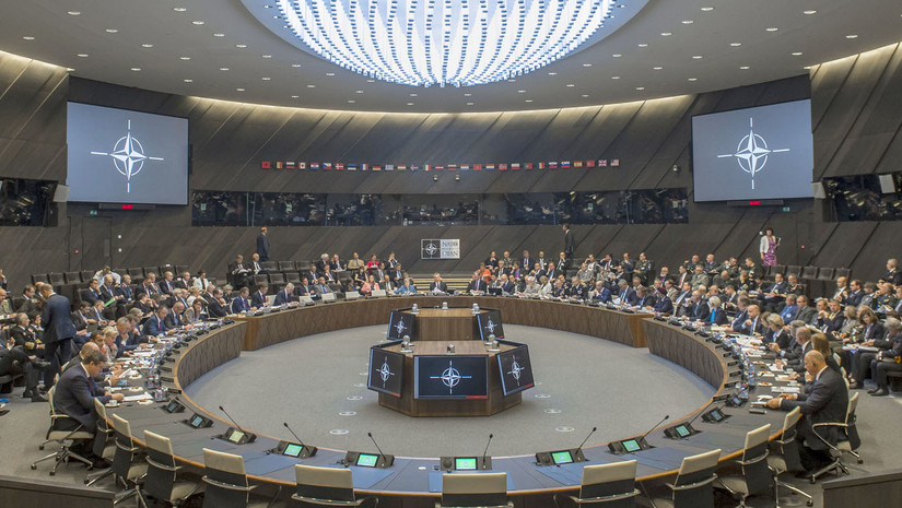 Defense ministers meet at the NATO headquarters in Brussels. A large conference room features a round table in the center of the image. More than 100 people are present.