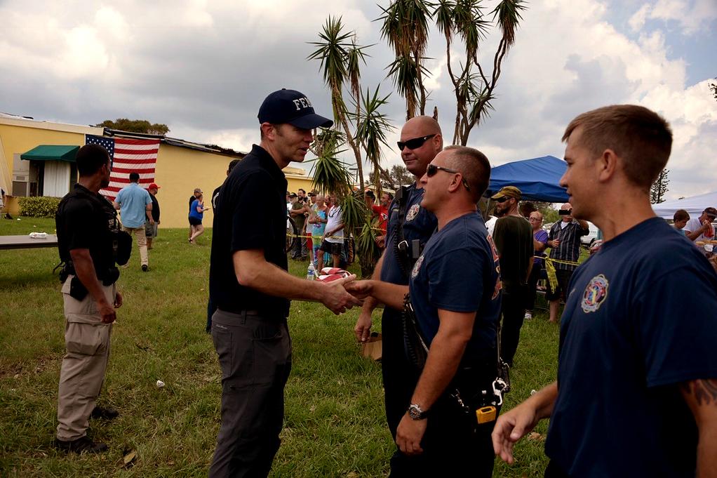 Brock Long shakes hands with a first responder. Behind him a single-wide trailer sits with an American Flag hanging. The sky is cloudy.