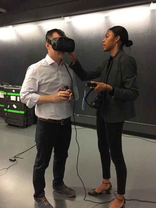 On the left, a man is standing wearing virtual reality goggles talking to a woman on the right about how to participate in the virtual reality world.