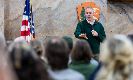 Secretary Zinke holds a microphone and talks to a group of people sitting inside.