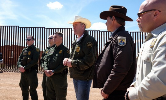 Secretary Zinke wears a cowboy hat and stands in a line with border patrol officers in front of a tall metal wall.