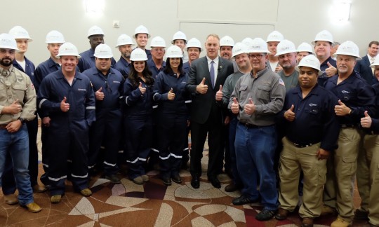 Secretary Zinke poses with a large group of workers in hard hats and coveralls in a conference room.
