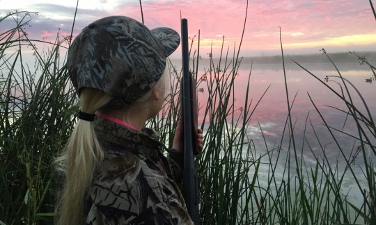 A young girl with blonde hair wears hunting camouflage and holds a shotgun while standing in tall grass and looking at a pink sunrise.