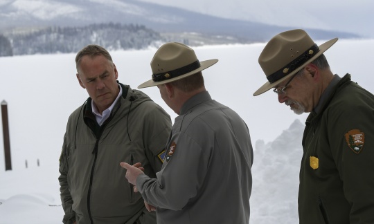 Two man wearing campaign hats talk with Secretary Zinke, snow and mountains in background