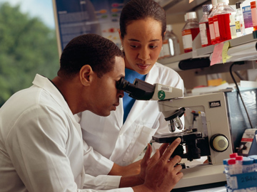 An African American male researcher looks through a microscope as Asian female researcher observes.