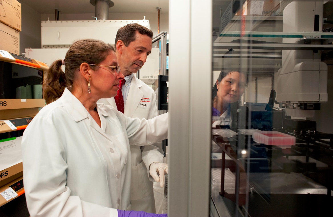 Three scientists in lab coats examine cell cultures in a large piece of lab equipment.