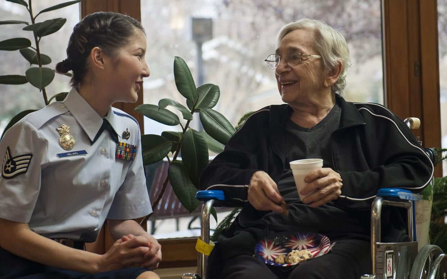 A female Servicemember with a smile is talking to a female Veteran who holds a cup of coffee sits on a wheelchair.