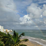 View from atop a building looks down on a sandy beach with a rainbow, palm tree just below view, and buildings in distance.