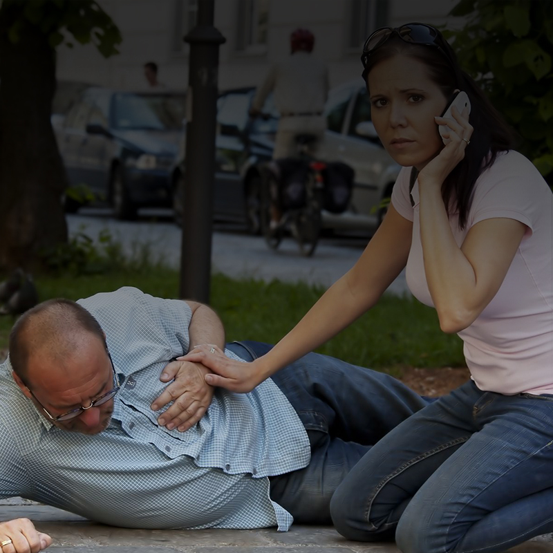 Photo of a man lying on the ground having a heart attack, while a woman on her cell phone tries to help him.