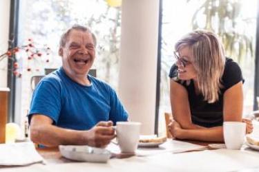 Older Caucasian man with Down syndrome, wearing a blue t-shirt, sits at a table with his right hand holding a white mug and a plate of food in front of him. He is laughing while a younger Caucasian woman with blonde hair and glasses and sitting to his left leans forward on the table and looks on, smiling.