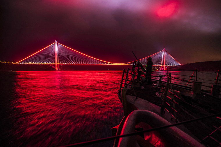 Sailors man a gun on a ship and look out over a suspension bridge illuminated in red light at night illuminated in red light.