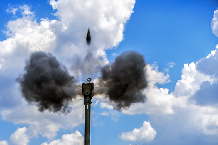 A howitzer round shoots into the blue sky as two gray puffs of smoke burst from the weapon's muzzle.