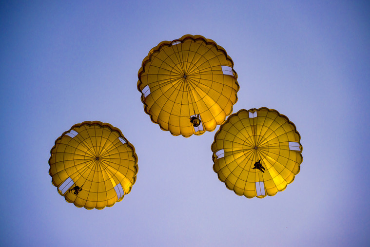 Three yellowish parachutes, shown from below, descend in a blue sky.