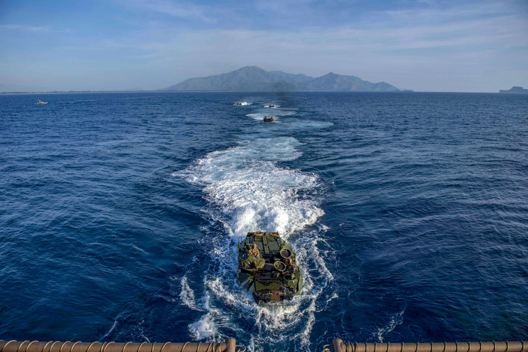 Amphibious military vehicles make their way toward a ship in the sea.