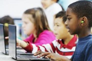This photo shows three young elementary-aged children in front of laptops in a classroom. The child in the forefront, in focus, in the photo, is an African-American boy wearing a navy blue pocketed t-shirt and wearing neon green earbuds plugged into the laptop. He is typing at the keyboard. To his right is a young boy wearing a white and red striped collared shirt, also at a laptop, and to his right is a young Caucasian girl in a bright pink long-sleeve shirt, also at a laptop.
