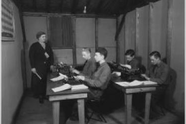 This vintage black and white photo displays a typing class with two rows of tables with two male students at each table with typewriters in front of them. A female instructor is at the front of the classroom with a ruler in her hand. 