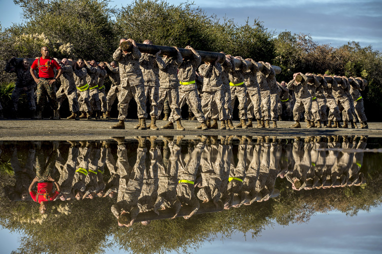 Marines carry logs on their shoulders outside by a body of water, which shows their reflection.