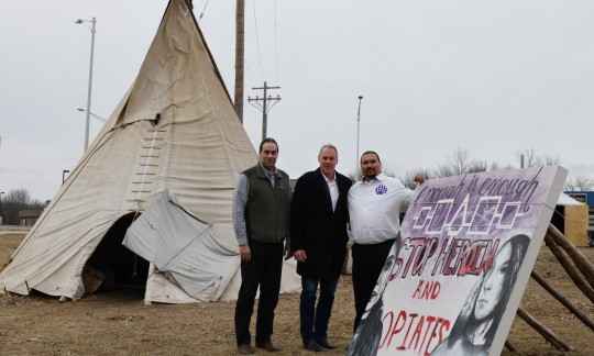 Secretary Zinke stands outside with two Native American men next to a large canvas tipi and a mural calling for an end to heroin and opioid abuse.