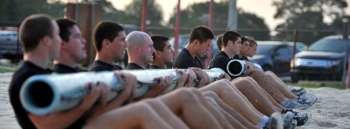 A military training shows Servicermembers sit side-by-side on the sand ground and lift the metal pipes with their arms. 