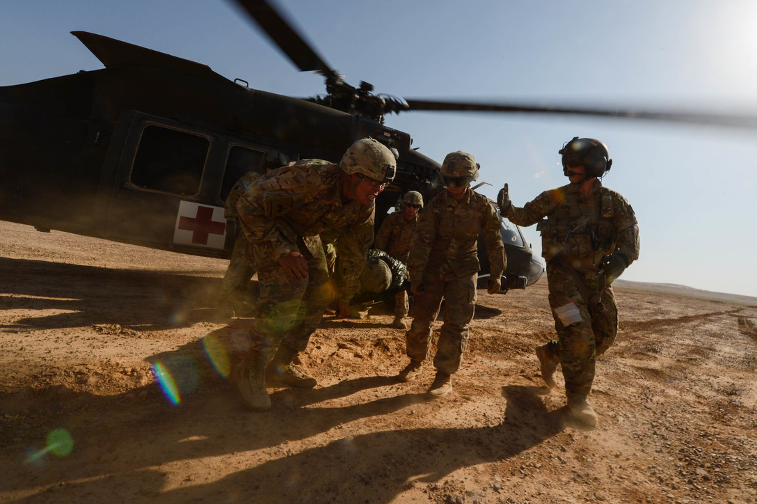 Soldiers run away from a helicopter carrying a stretcher on desert-type terrain.