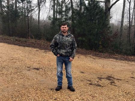 A man wearing a camouflage hoodie posing for a photo on a gravel road