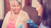 Caring, Latin descent home healthcare nurse gives vaccine or medicine injection to African descent senior adult patient at home, assisted living, or nursing home setting. She holds the syringe and wears protective gloves. The female patient happily receives the shot while sitting on her comfy sofa. Kitchen background.