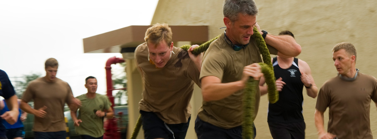 A group of male soldiers are running and two of them are pulling a thick rope from their shoulders.