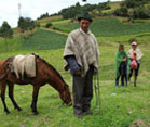 In lush green mountains of Colombia, a man looks at the camera holding the reigns of a horse, women in background