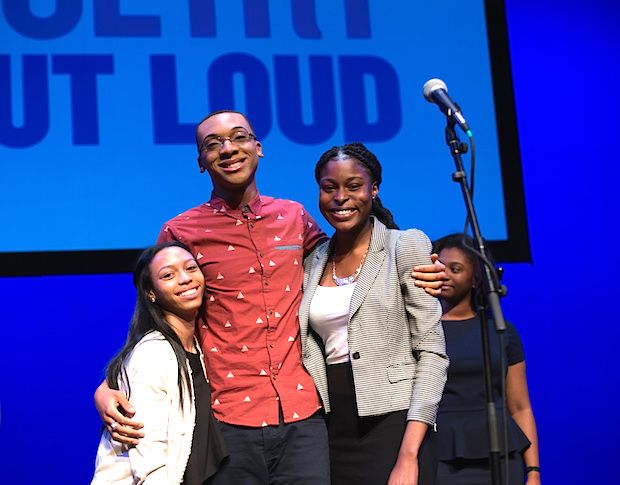 Two young woman and a young man stand together on a stage with other students behind them.