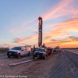 Image shows a drill rig and vehicles near a road at dawn