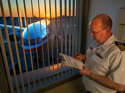 Airport offical reviews a checklist on a clipboard next to a window. Outside the window there is commercial airplane in front of a setting sun.
