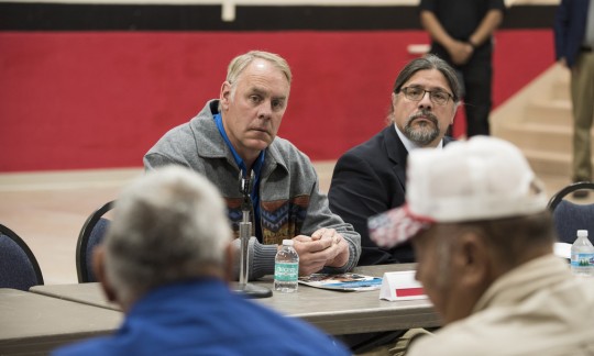 Secretary Zinke sits at a table in a large room talking to a group of people.