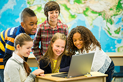Foto de estudiantes mirando información en la computadora portátil en un salón de clase que tiene un mapa en la pared.