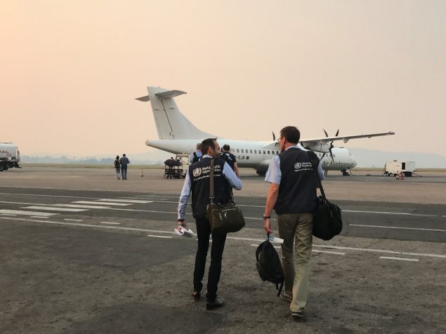 Two men from the back representing the World Health Organization. The two men in the foreground are carrying travel bags, wearing blue WHO vests, and walking across the tarmac towards a prop plane. There are four other people and mountains with a hazy skyline in the distance.