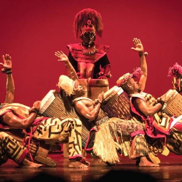 African drummers onstage in traditional costume leaning back in a dance pose with a tall man behind against a red background