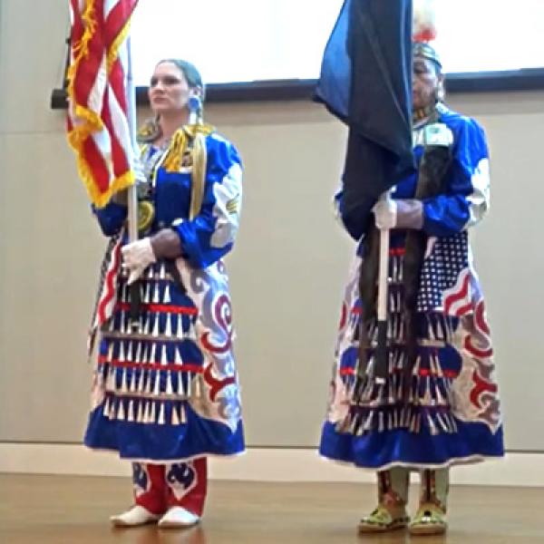 Two women in native american traditional dress on a stage, one holding a U.S. flag and the other holding a tribal flag