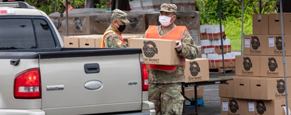 [Photo: A member of the National Guard loads a food box onto a resident's truck]. 