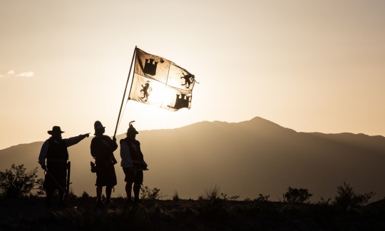 Three men dressed as Spanish explorers wave a flag while standing on a hillside in bright sunlight.