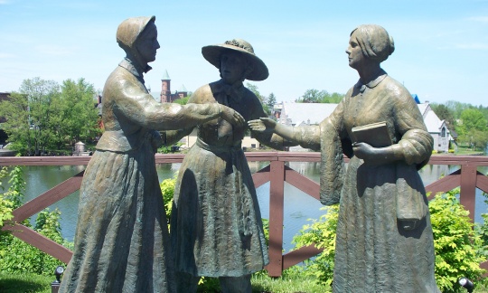 Statues of 3 women against a blue sky; women in hat in a pose introducing woman with no hat carrying book, to a woman in a bonnet