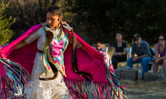 A young woman in traditional tribal clothing dances outside as a small group of people look on.
