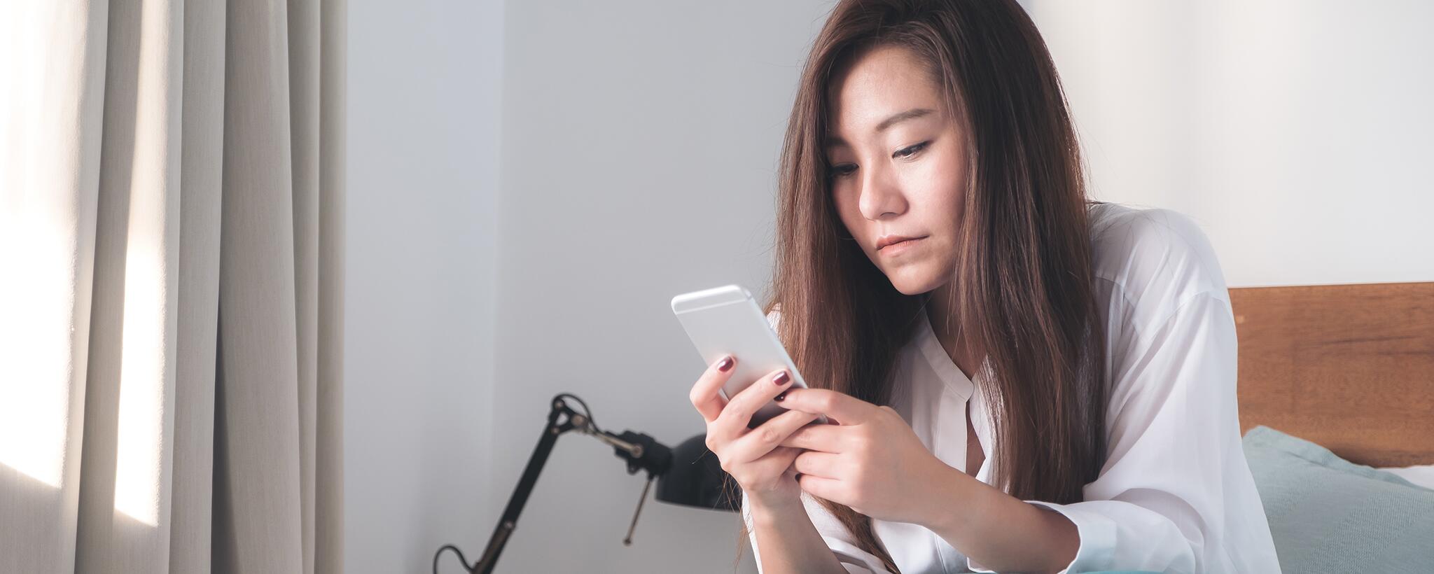 Una mujer sentada en su cama mirando su teléfono inteligente