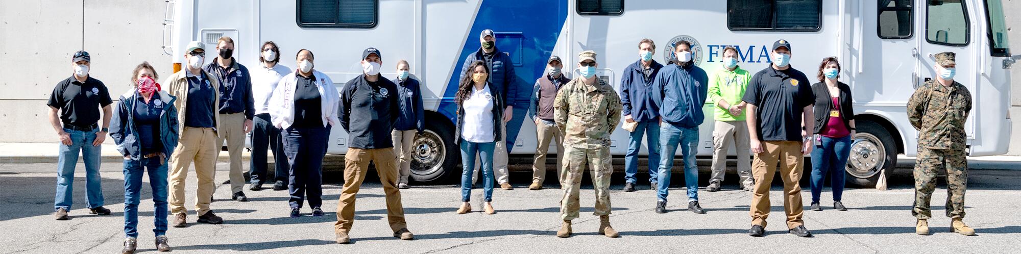 multiple men and women standing in front of a fema trailer with mask on and their hands behind their back. 