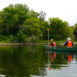 ​USGS employee in a canoe hold a prism while survey rod level and the channel point is collected with a total station from shore