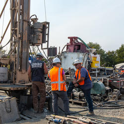 Image shows three men in safety equipment standing next to a drill rig