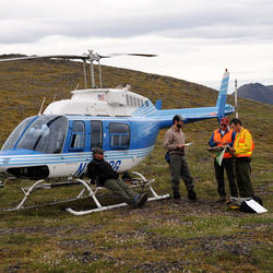 Image shows a helicopter with four USGS scientists talking to each other next to it, with grasslands in the background