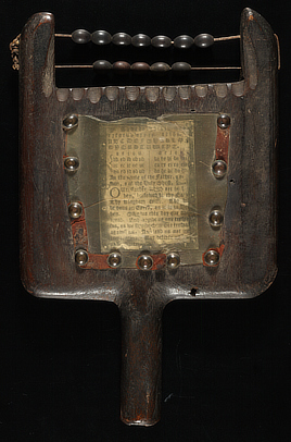 An antique wooden tablet  with a h handle at the bottom holds a small sheet of paper covered by a transparent sheet of horn. The alphabet and Lord's Prayer are printed on the paper. Along the top edge are small rails with abacus beads for doing arithmetic.  
