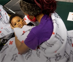 woman holds child in red cross shelter, wrapped in a red cross blanket