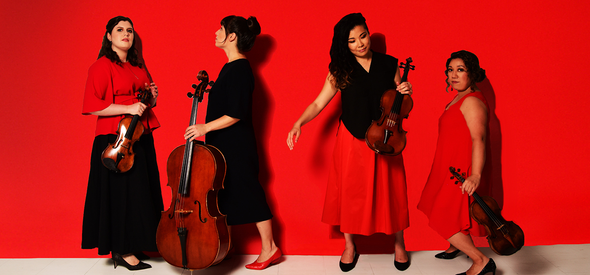 Four women holding stringed instruments facing various directions and standing in front of a vivid red background