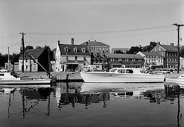 Long boats are moored in a waterway near a road lined with commercial buildings and parked cars.